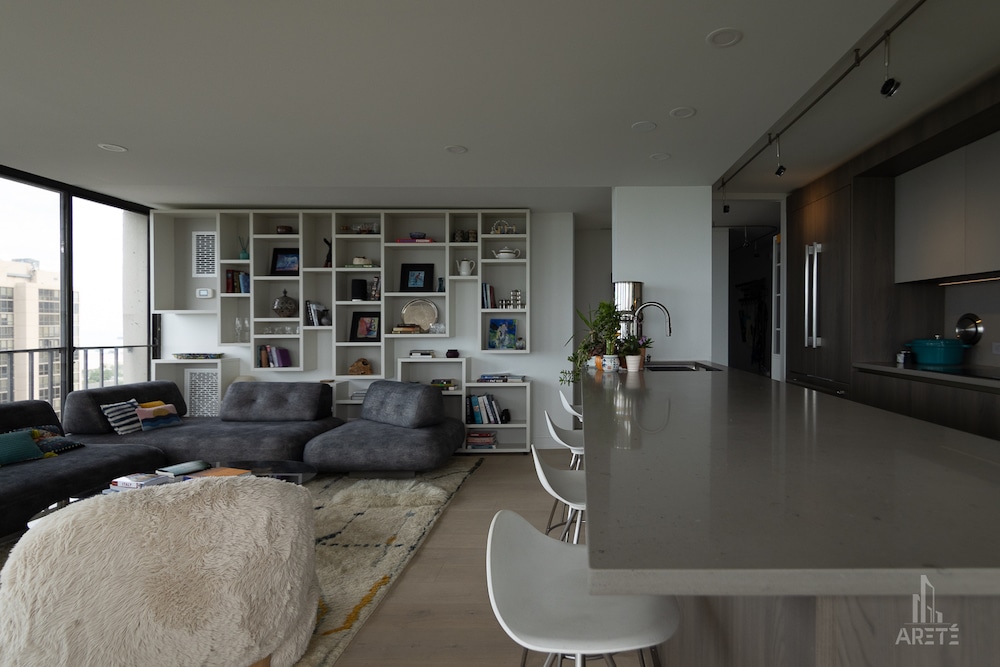 A contemporary open-concept living room featuring a large custom white shelving unit filled with decor, a dark grey sectional sofa, a fuzzy white ottoman, and floor-to-ceiling windows, viewed from behind a kitchen island.