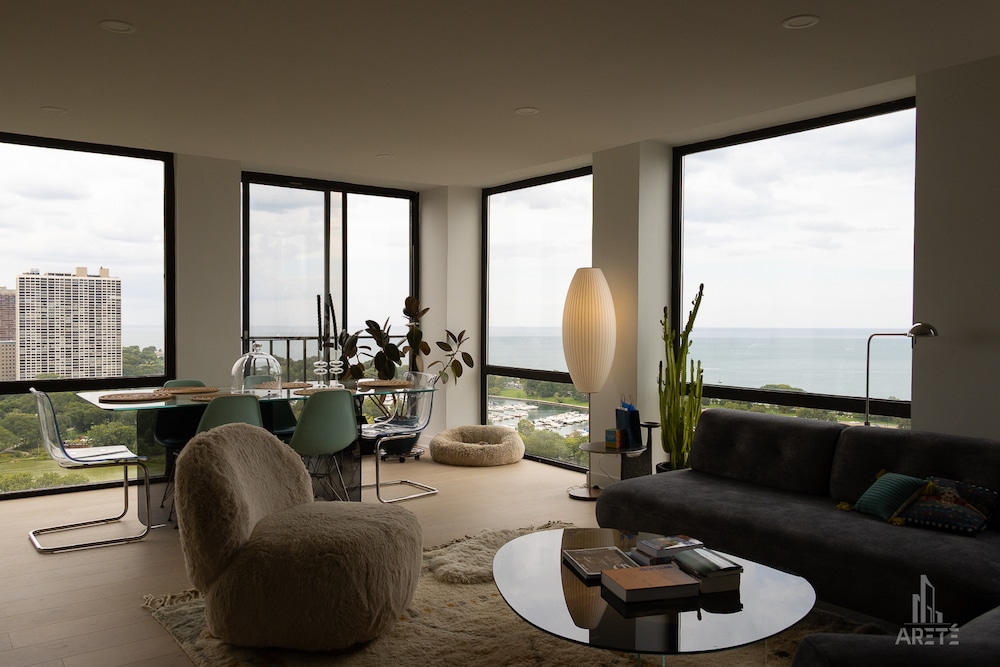 A bright condo living and dining area featuring floor-to-ceiling corner windows with a view of a harbor and lake, furnished with a glass dining table, a fuzzy accent chair, a dark grey sofa, and a tall cactus.