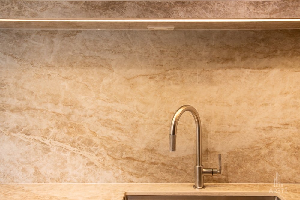 Modern stainless steel kitchen faucet with curved spout over beige marble backsplash and counter.