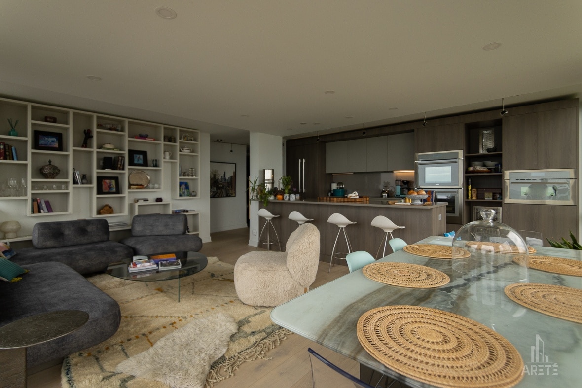 Areté_open concept - Arete' Open-concept living space with dark gray sectional sofa, glass coffee table, and a cream shag rug, adjacent to a modern kitchen with an island and white bar stools.