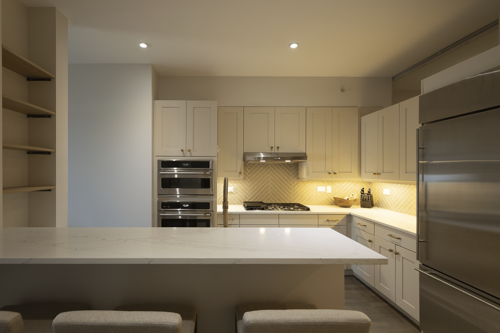 Bright modern kitchen with white cabinets, marble island, and stainless steel fridge and wall ovens in a well-lit space.