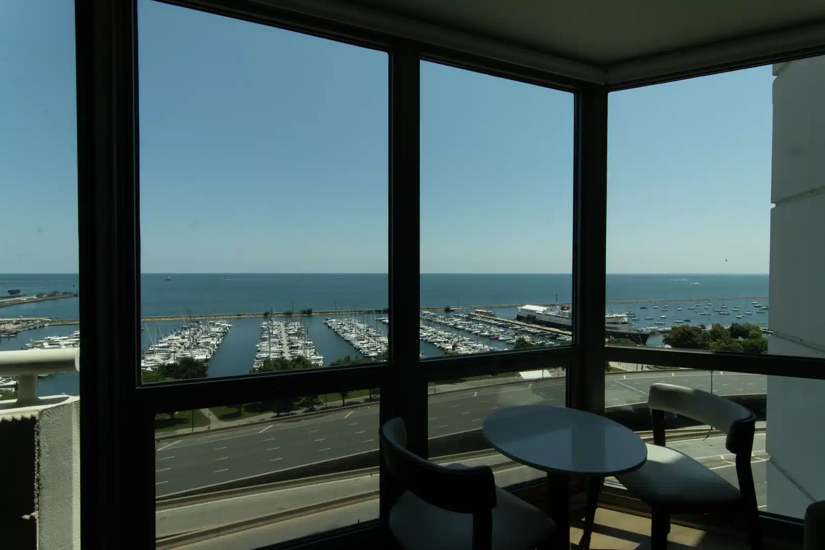 Indoor balcony seating with a round table and chairs, overlooking a marina filled with boats and the blue sea beyond.